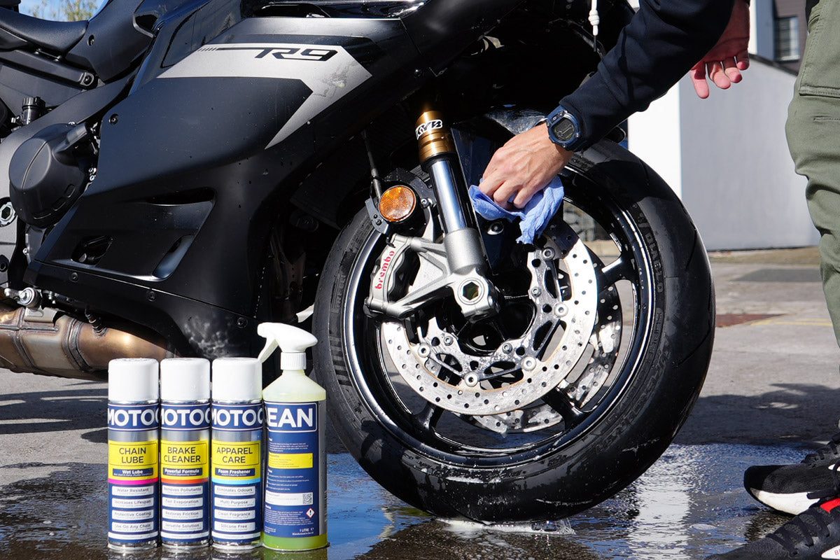 Person cleaning a motorcycle front wheel during a pre-ride check, with MotoClean products beside the bike.