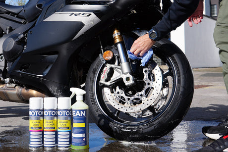 Person cleaning a motorcycle front wheel during a pre-ride check, with MotoClean products beside the bike.